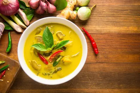 Aromaticzne curry z tofu, podane w białej misce, otoczone świeżymi składnikami: chili, trawą cytrynową, imbirem i cebulą.