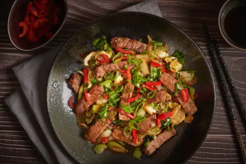 Pyszne wieprzowina w sosie teriyaki z papryką i szczypiorkiem, podana na ciemnym talerzu.