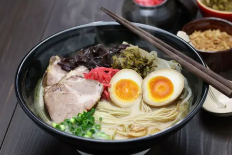 Parujący makaron do ramen z plastrami wieprzowiny, jajkiem na pół, szczypiorkiem i marynowanymi dodatkami.