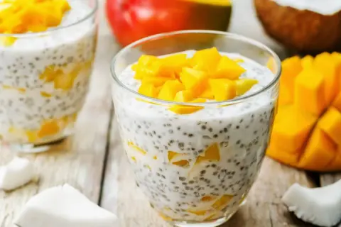 Pyszny deser chia z mlekiem kokosowym i kawałkami mango, podany w szklance. Obok świeże mango i wiórki kokosowe.