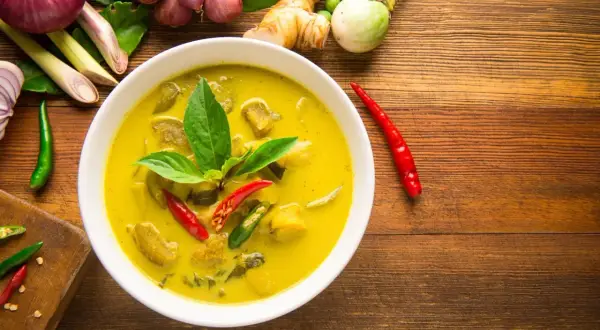 Aromaticzne curry z tofu, podane w białej misce, otoczone świeżymi składnikami: chili, trawą cytrynową, imbirem i cebulą.