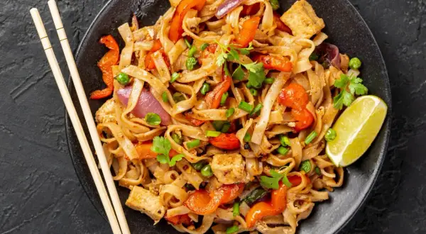 Pyszny kurczak pad thai z warzywami i tofu, podany z limonką i pałeczkami.