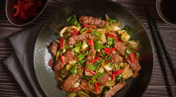 Pyszne wieprzowina w sosie teriyaki z papryką i szczypiorkiem, podana na ciemnym talerzu.