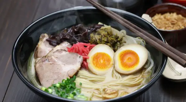 Parujący makaron do ramen z plastrami wieprzowiny, jajkiem na pół, szczypiorkiem i marynowanymi dodatkami.