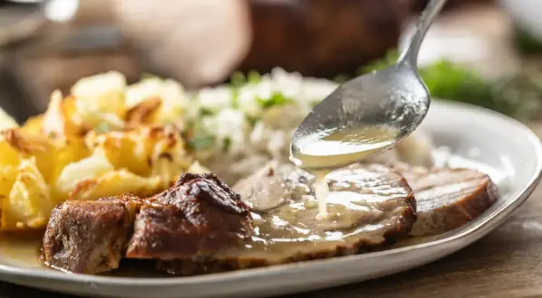 Łyżka polewa pieczoną karkówkę aromatycznym sosem. Obok ryż i złociste ziemniaki.