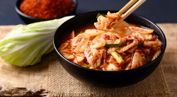 Miseczka z kimchi, pałeczki podnoszące kawałek kapusty, liść kapusty i miseczka z przyprawą.