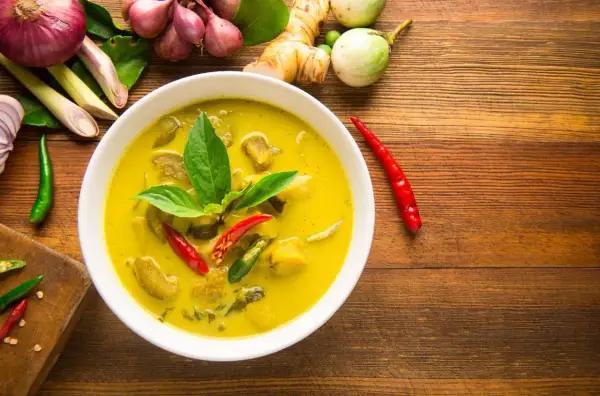 Aromaticzne curry z tofu, podane w białej misce, otoczone świeżymi składnikami: chili, trawą cytrynową, imbirem i cebulą.