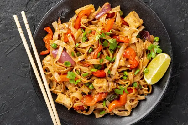 Pyszny kurczak pad thai z warzywami i tofu, podany z limonką i pałeczkami.