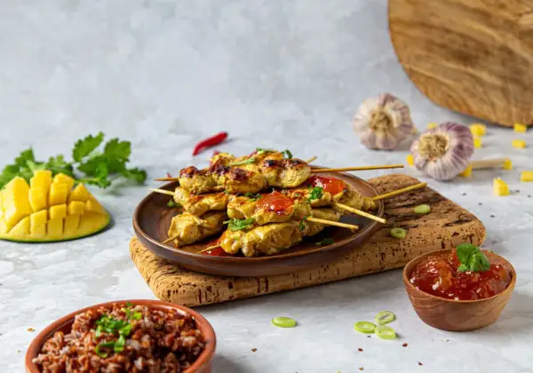 Aksamitne satay z kurczaka na patyczkach, polane sosem, obok ryż, mango i czosnek.