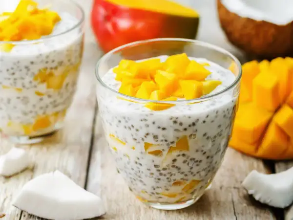 Pyszny deser chia z mlekiem kokosowym i kawałkami mango, podany w szklance. Obok świeże mango i wiórki kokosowe.